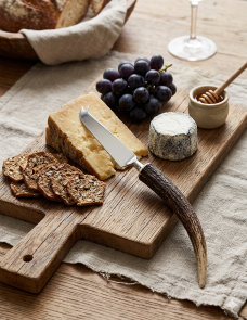 An antler handle cheese knife rests on a rustic wooden board next to a selection of cheeses, wine glasses in the background.